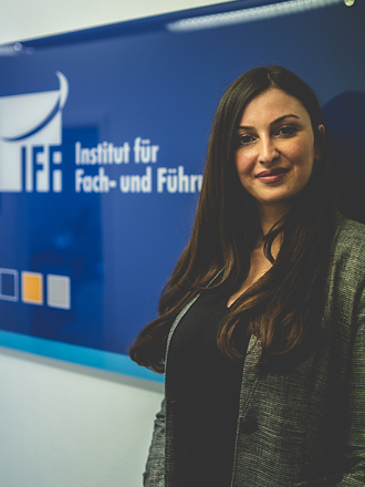 Eine Frau mit langen dunklen Haaren und einem grauen Blazer steht vor einem blauen Schild mit der Aufschrift Institut für Fach- und Führungskräfte. IFF Akademie - kaufmännische Weiterbildung