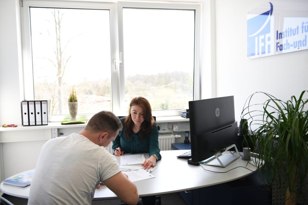 Eine Frau sitzt mit einem Mann an einem Schreibtisch, beide sehen sich Dokumente an. Sie befinden sich in einem hellen Büro mit einem großen Fenster, Pflanzen, einem Computer und einem Schild an der Wand, auf dem "Institut für Fach-und. IFF Akademie - kaufmännische Weiterbildung